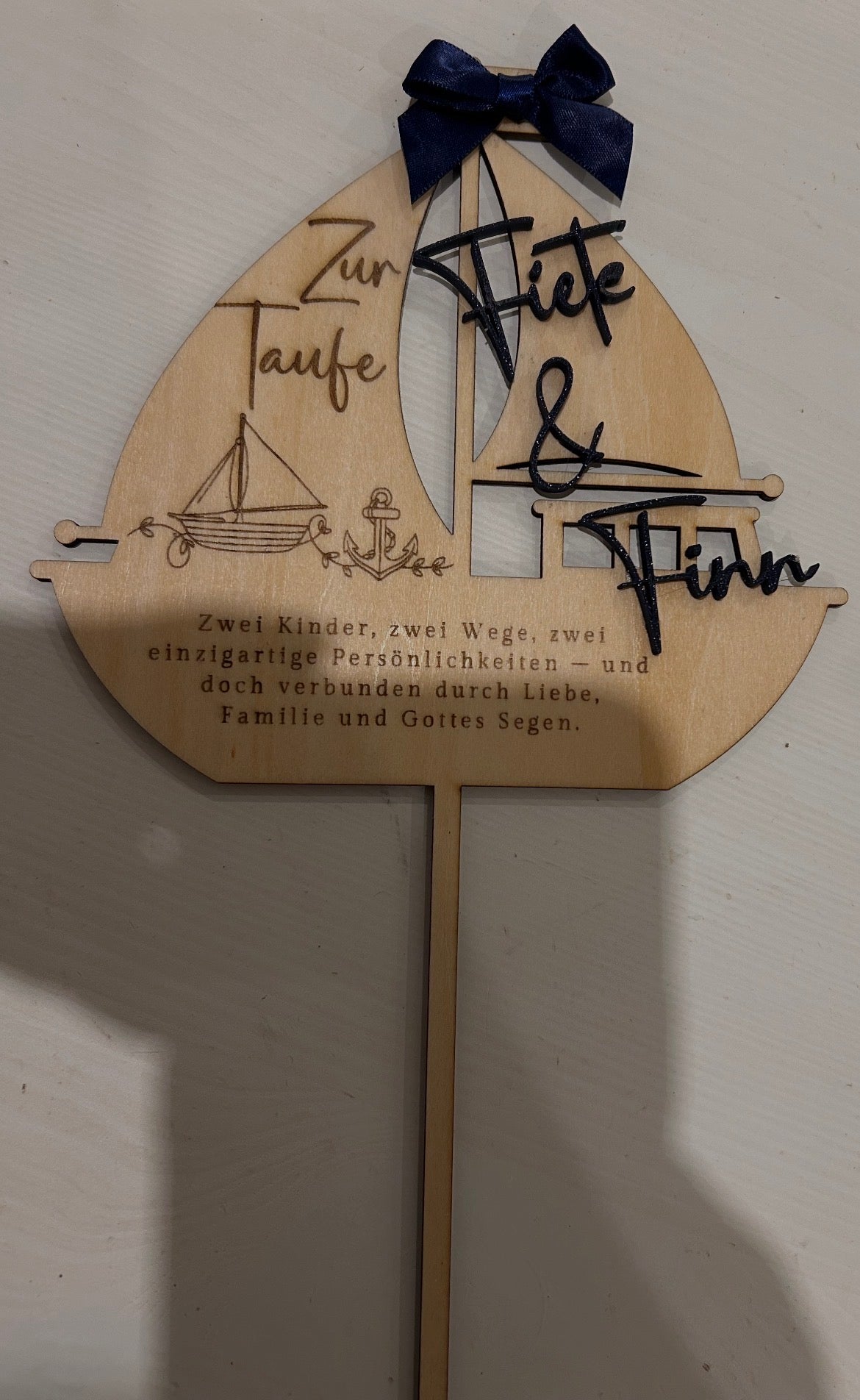 Caketopper aus Holz in Segelbootform zur Taufe mit zwei aufgesetzten Namen und dunkelblauer Schleife.